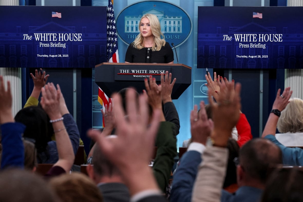 A woman stands at a lectern in the White House while reporters raise hands to ask questions.