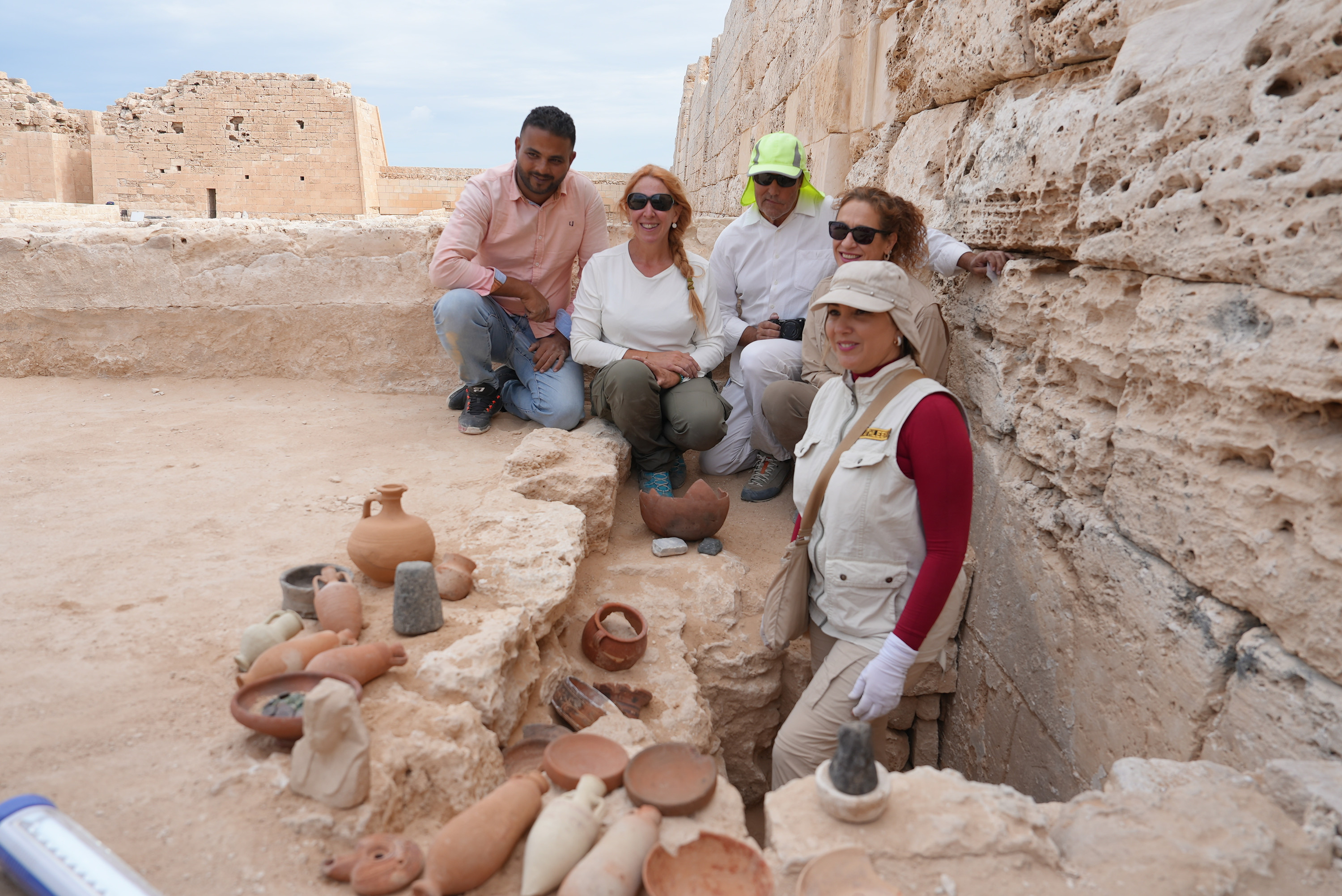 Archaeologists pose at a sandy excavation site with ceramics and coins.