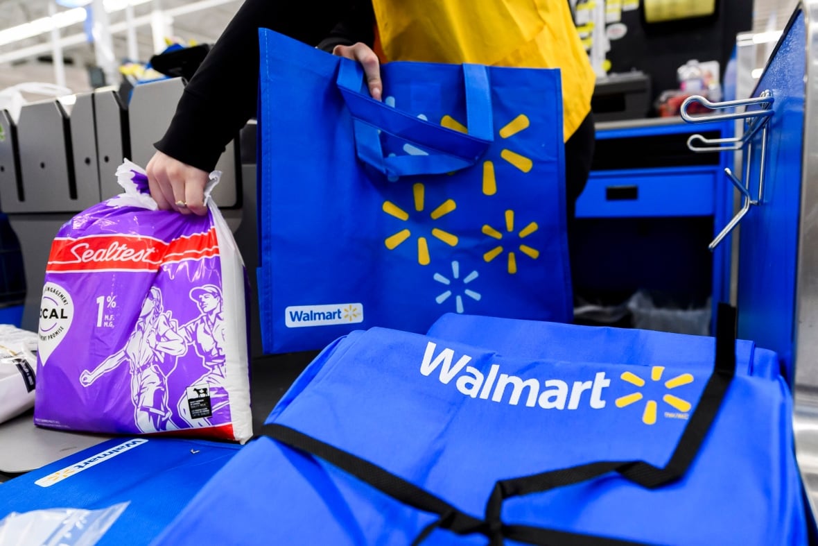 A Walmart employee in a yellow vest grabs a bag of milk to place it into a blue Walmart branded reusable bag at a checkout. 