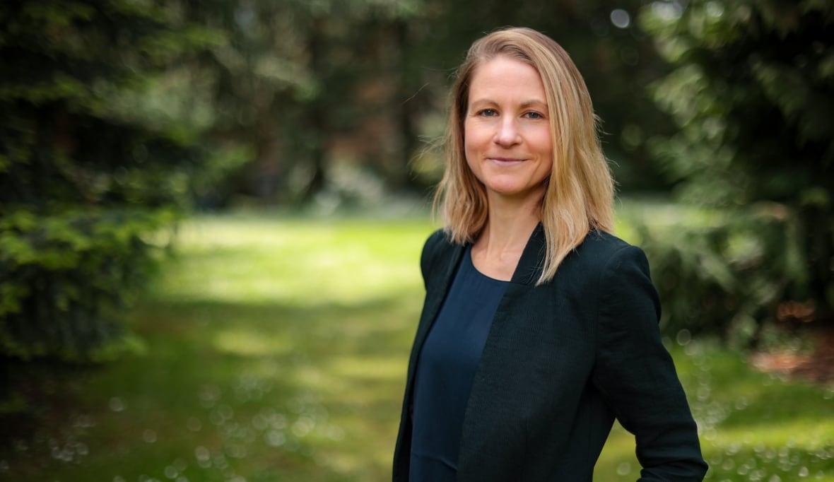 A woman in a dark coloured blazer and blonde shoulder length hair stands and smiles.