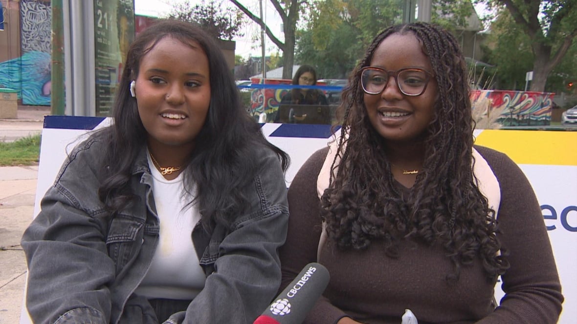 Two young female university students sit on a bench at a bus stop and smile.