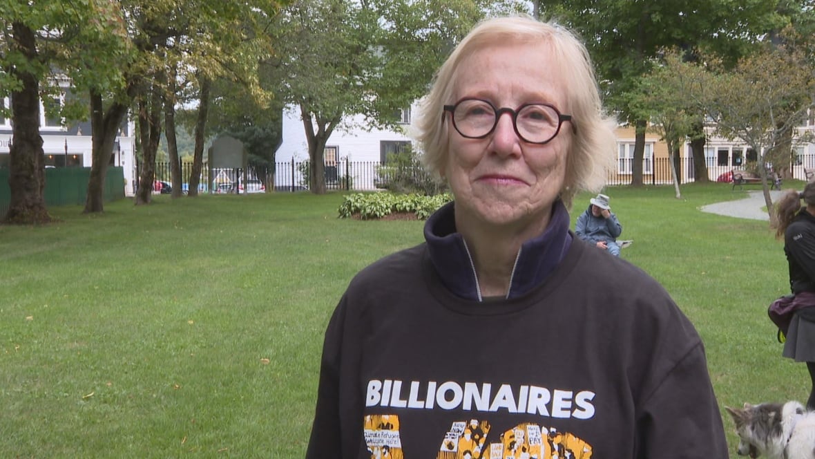 A woman with white hair and round classes smiles to the camera, standing in a park with green grass.