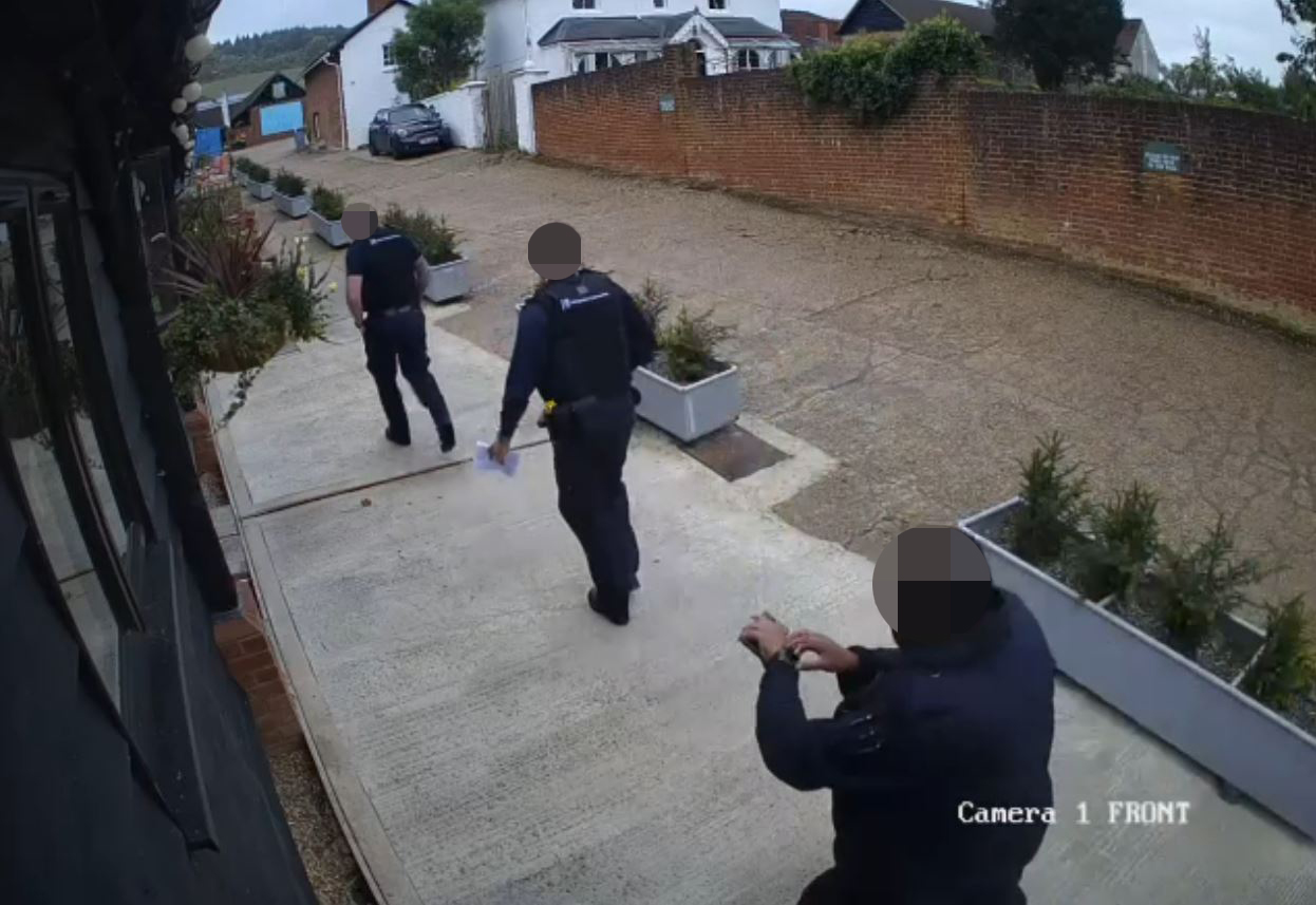 Two police officers walking away from a building, one holding papers, while a third person in the foreground records.