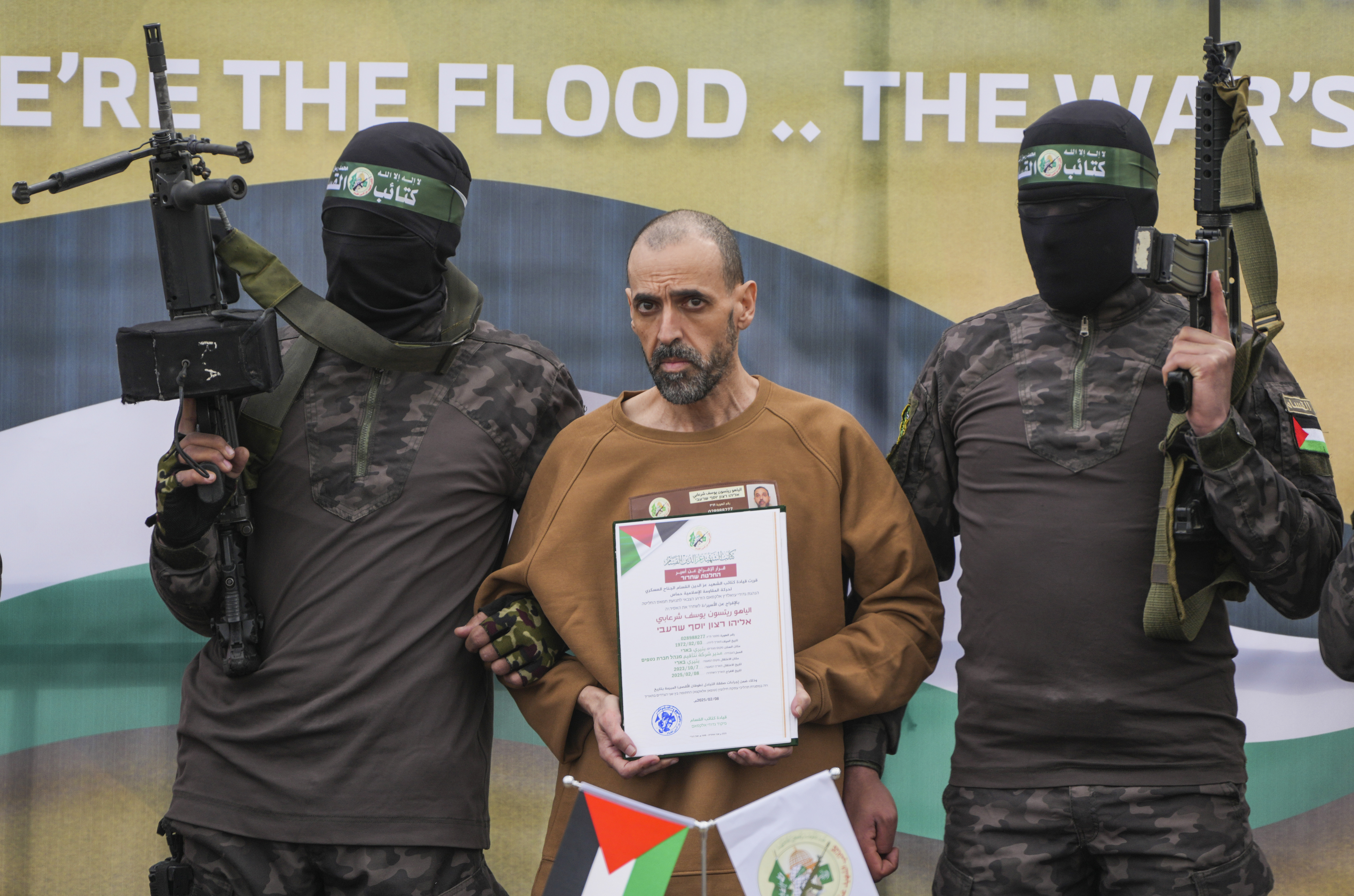 Eli Sharabi, an Israeli captive, escorted by two masked Hamas fighters with weapons, holding a document in Arabic and Hebrew with "Decision to release a prisoner" written on it.