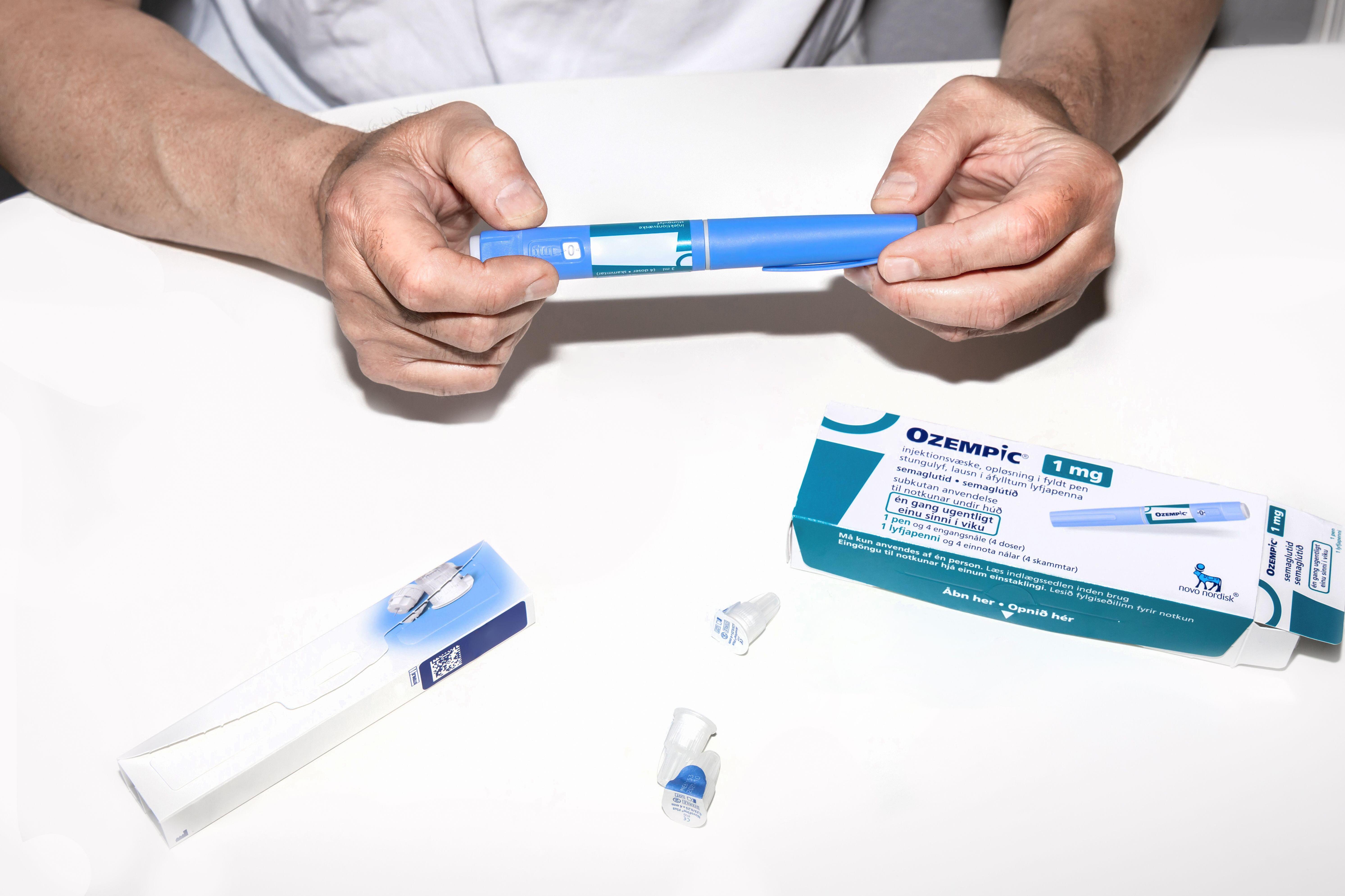 A person's hands holding an Ozempic insulin injection pen, with the Ozempic packaging and needles visible on a white surface.