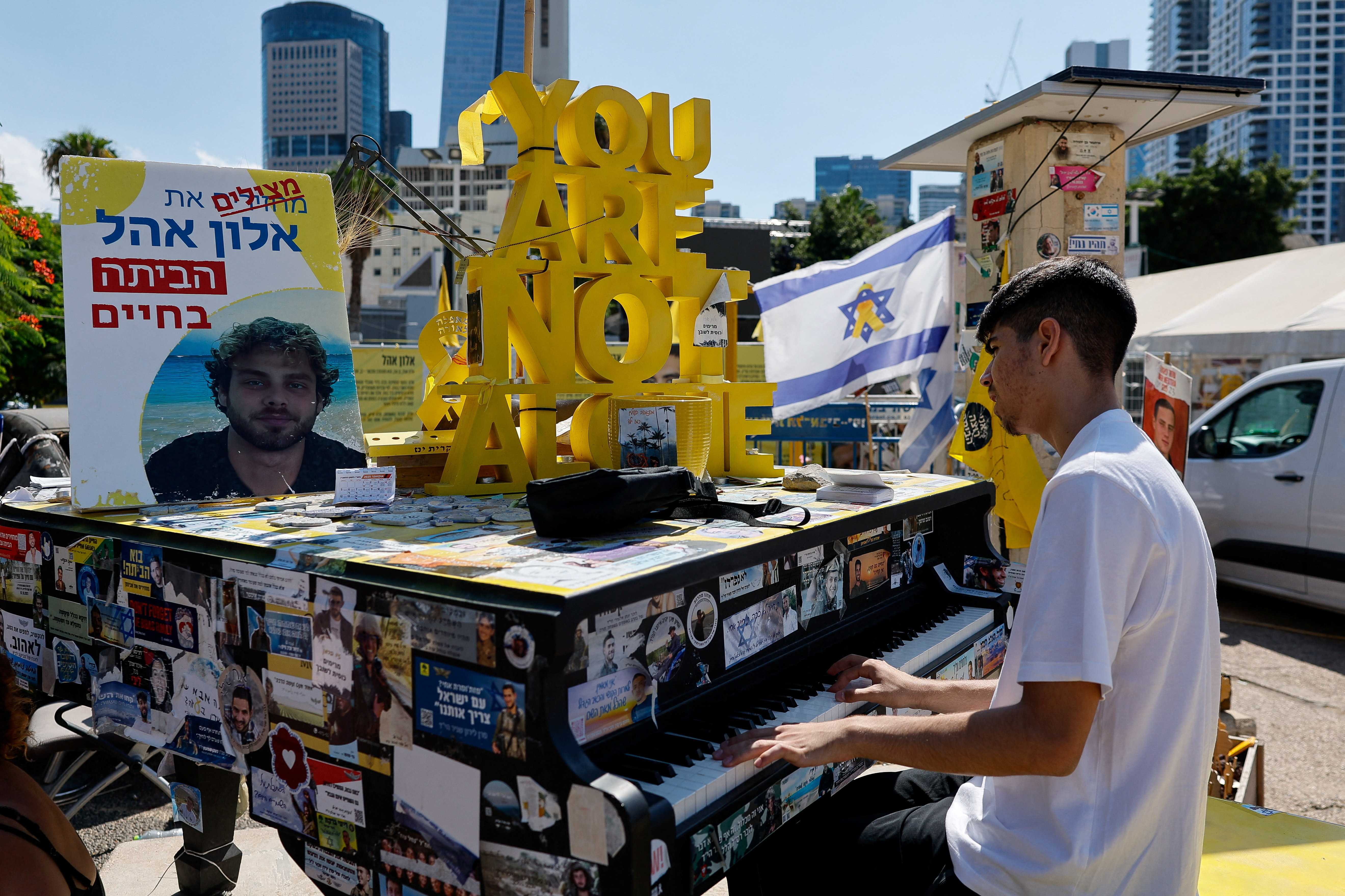 A boy plays the piano at "Hostages Square", with a sign reading "YOU ARE NOT ALONE" and an Israeli flag in the background.