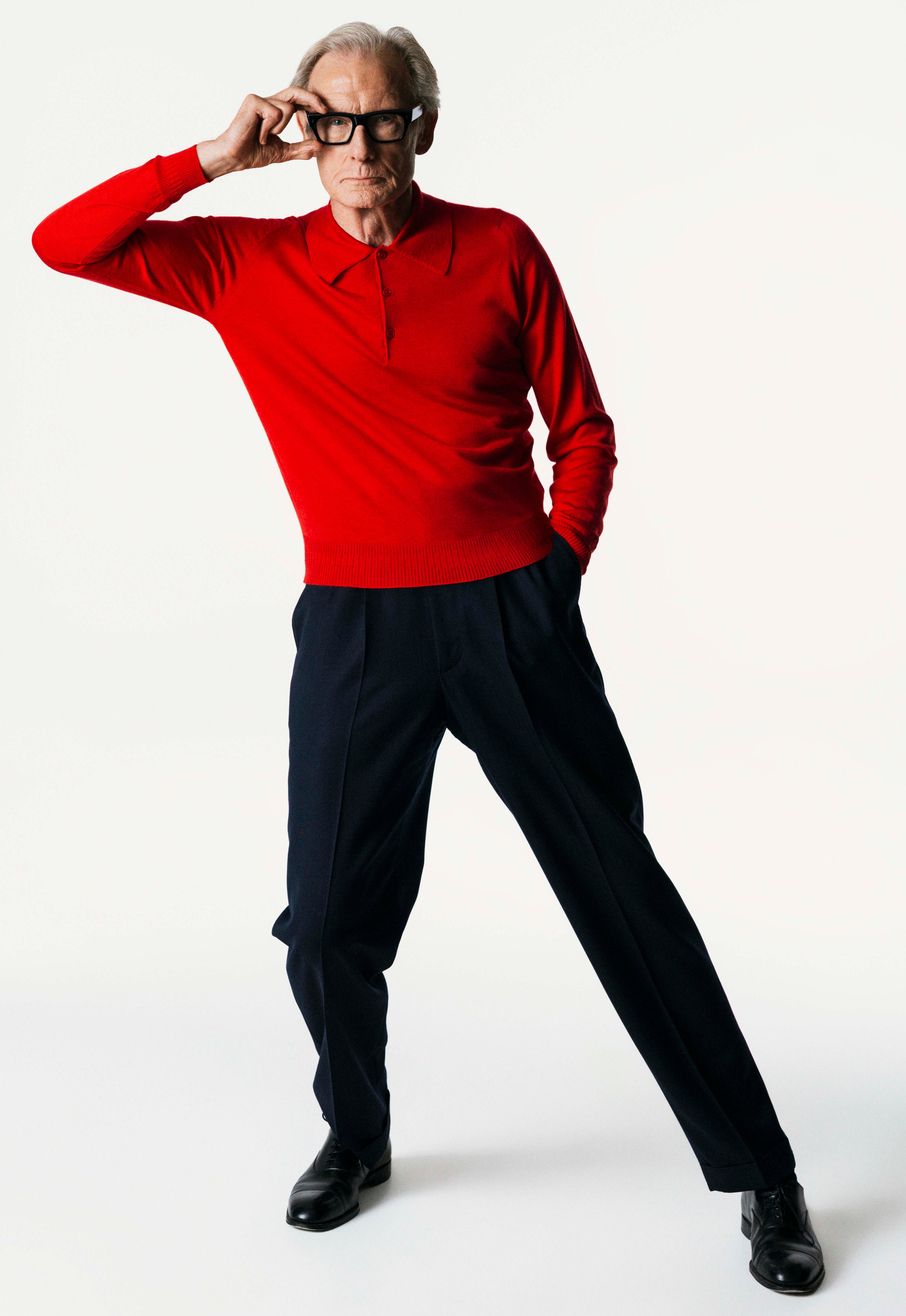 Bill Nighy in a red polo shirt, black trousers, and black shoes, holding his glasses up.