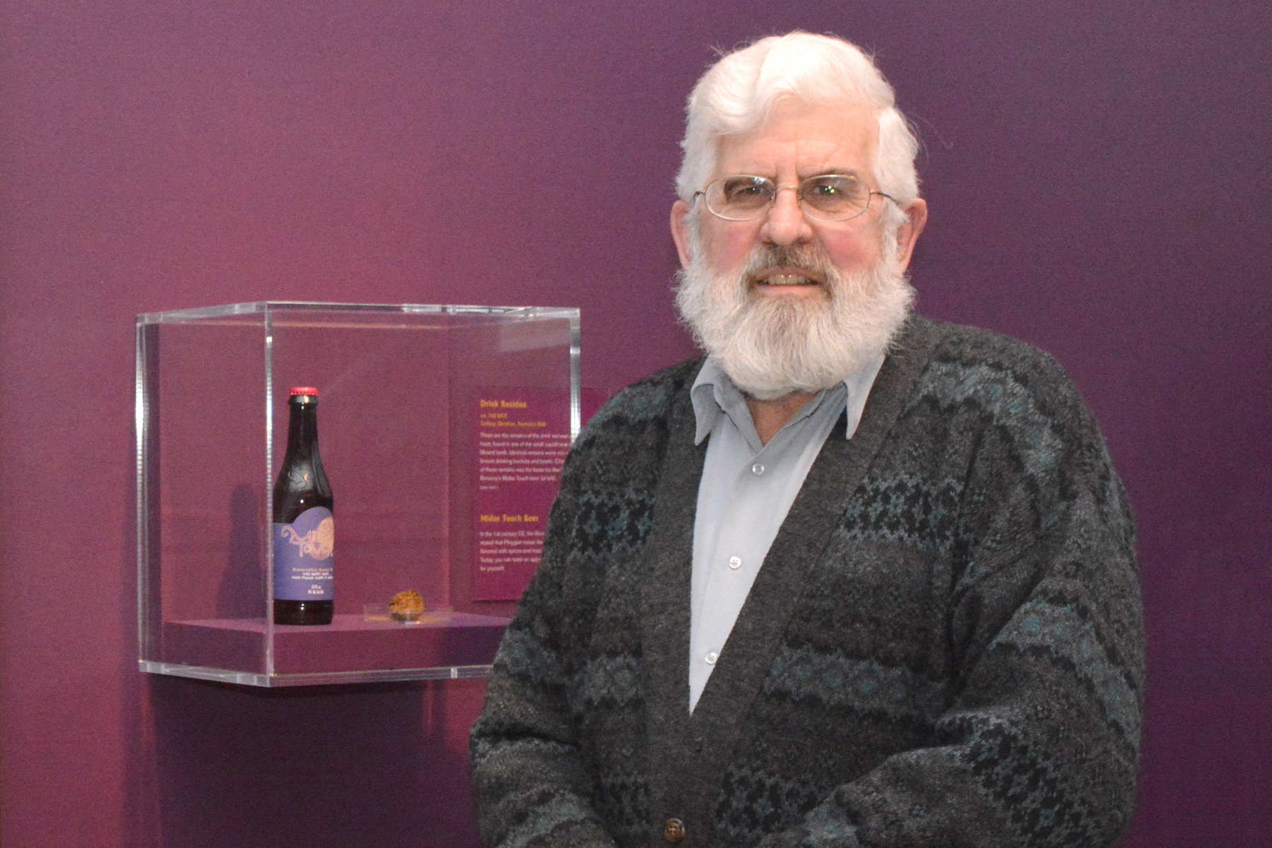 Dr. Patrick McGovern holding a bottle of Midas Touch brew, next to a display case in a museum.