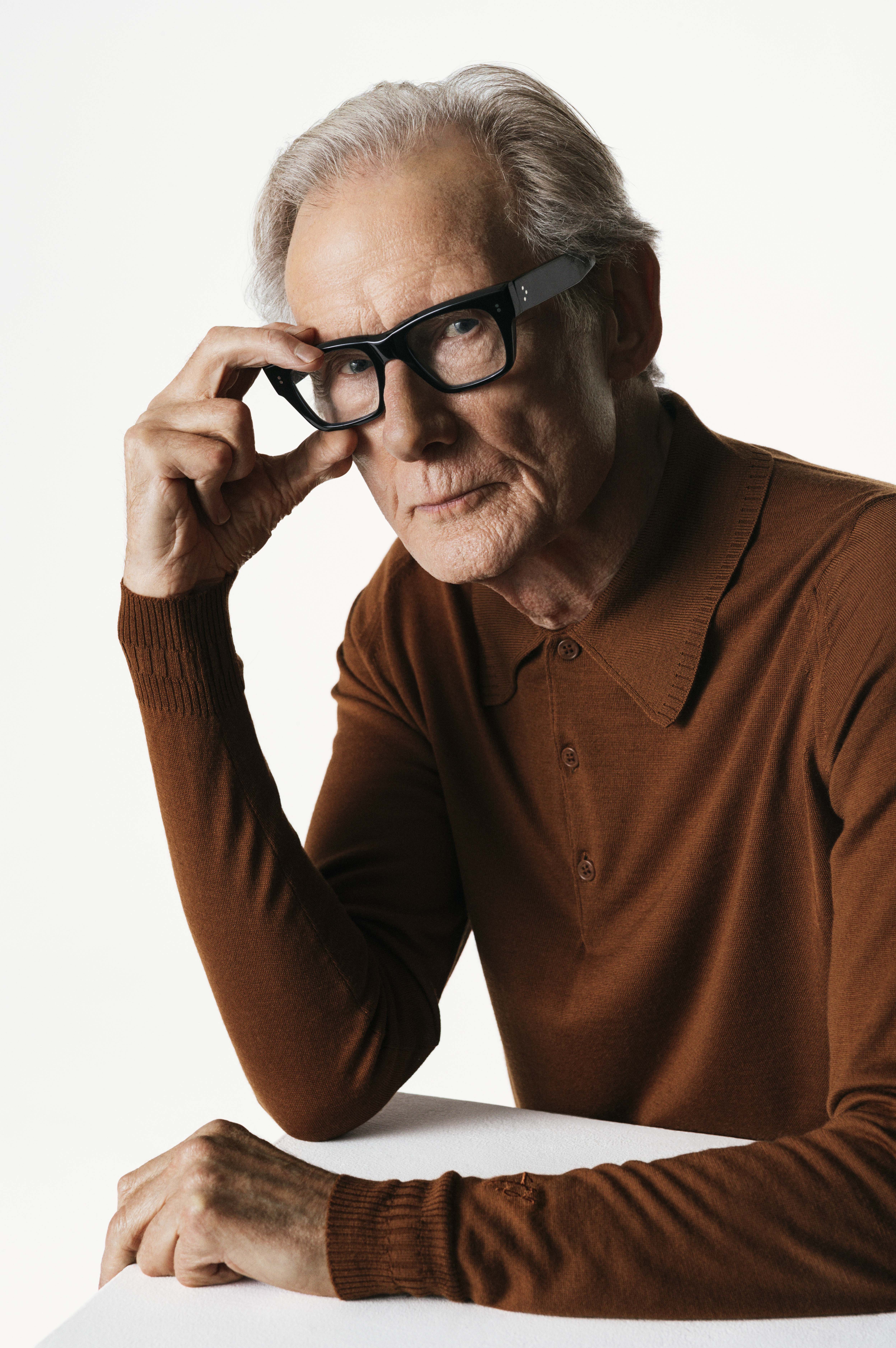 Bill Nighy wearing black glasses and a brown collared shirt.