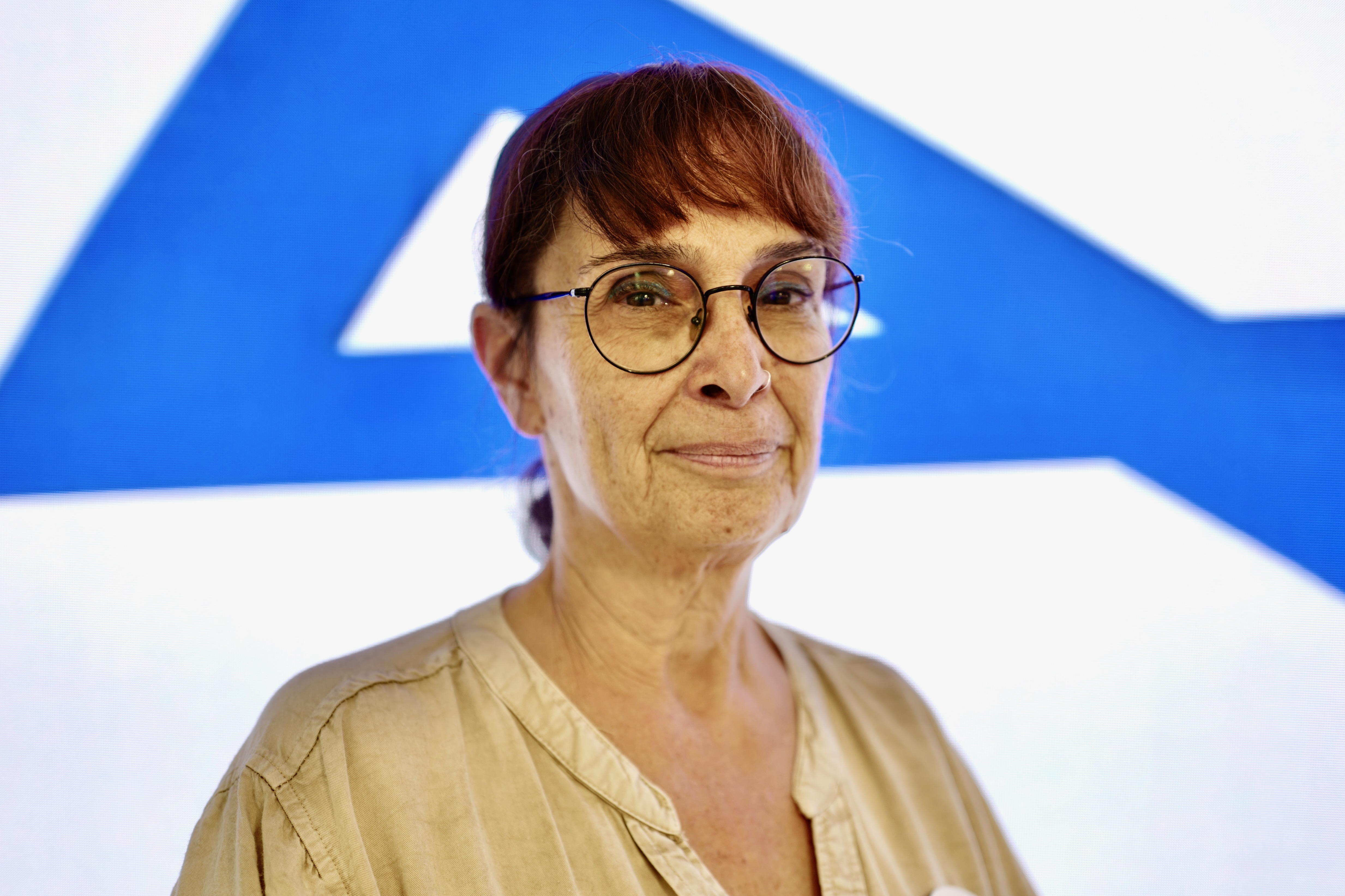 Head of nutrition Tamar Pfeffer-Gik, wearing glasses and a tan top, stands in front of a blurred blue and white background.