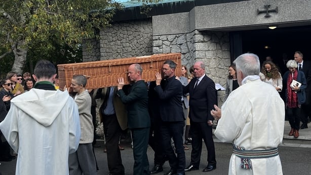 Men dressed in dark suits carry a brown coffin on their shoulders. Two priests wearing white robes are in front of them. A grey stone church is in the background.