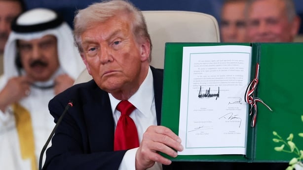 US President Donald Trump, wearing a white shirt, red tie and dark coloured jacket, holds up a white piece of paper inside a green folder.