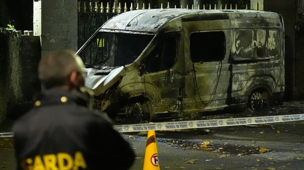 A man with 'garda' on his jacket looks at the remains of a burnt out van