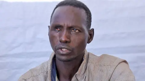 A head and shoulders shot of a man speaking. He is wearing a stripy, collared shirt. A blue cloth can be seen behind him.