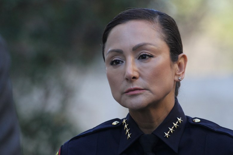 A person wearing a dark police uniform with gold stars on the collar stands outdoors, looking serious and focused. The background is softly blurred with natural light filtering through trees.