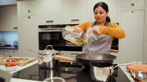 Getty Images A stock photo shows a woman pouring a big bottle of sunflower oil into a deep wok pan on an electric hob with cooking ingredients all around her and her kitchen cabinets and oven in the background.