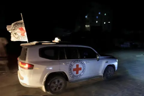 Reuters A white Red Cross vehicle with a cross emblem on its side and a flag waving from its roof, pictured on 27 October, 2025 against a night-time backdrop. 
