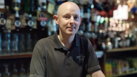 BBC Adam McClurg in a black t-shirt standing behind the bar in a pub. He is bald. There are spirits and drinks behind him.