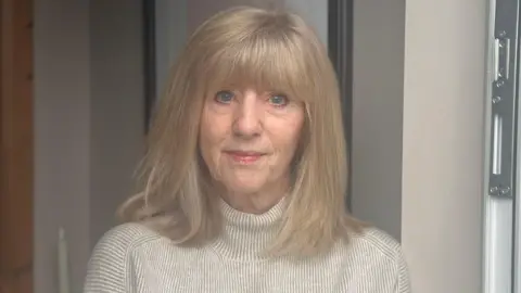 Janet Preston with long blonde hair and a beige polo neck standing at the front door of her house. There is a photoframe on the wall behind her.