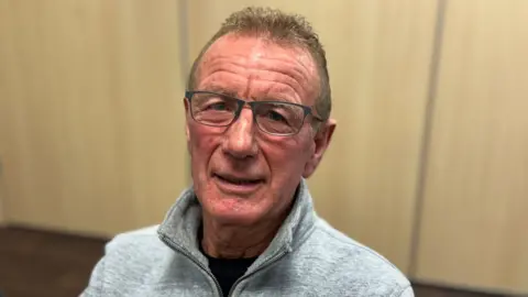 A man with short hair is looking into the camera. He is wearing a grey jumper with a zip and a black top underneath. He is wearing grey framed glasses. 