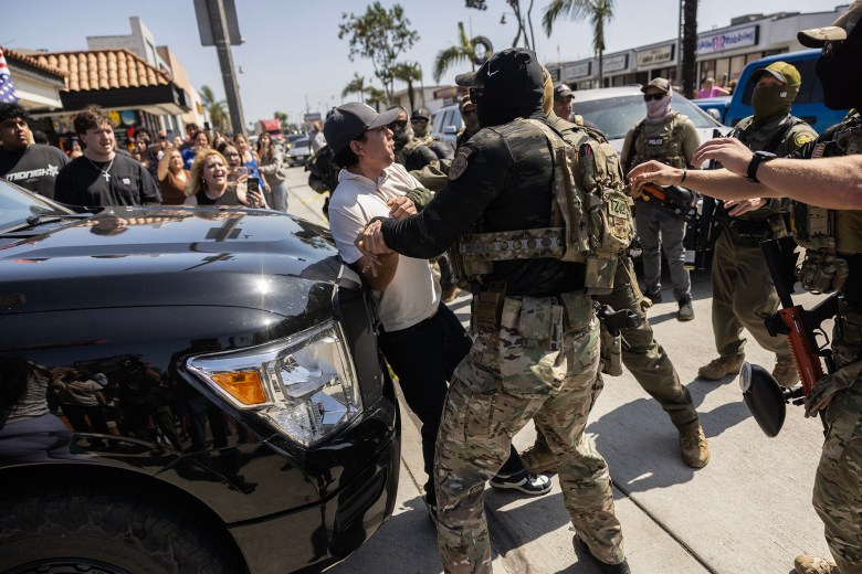 A tactical officer in camouflage grabs a person in a white T-shirt and gray cap, pressing them against a black pickup truck during a law enforcement raid. Several other officers in tactical gear and masks surround the scene, some carrying paintball-style weapons. In the background, a crowd of onlookers gathers on the sidewalk, some recording the event with their phones. The raid takes place on a busy commercial street lined with shops and palm trees.