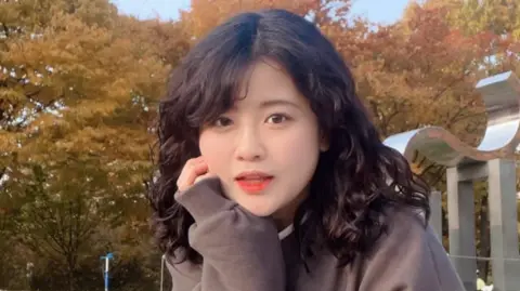 Instagram / Baek Se-hee Baek Se-hee resting her cheek on her hand as she looks at the camera. She has curly black hair and is wearing a brown sweater. In the background are trees with yellow leaves.