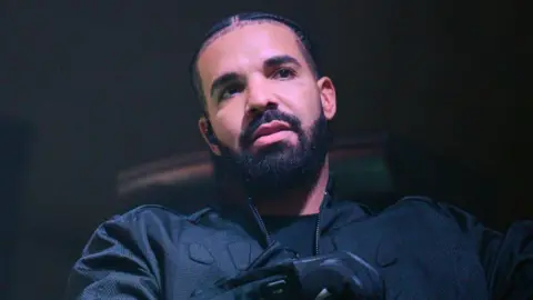 Getty Images Canadian rapper Drake is dressed in a dark shirt, jacket and gloves and holds a microphone while looking to off to his left.