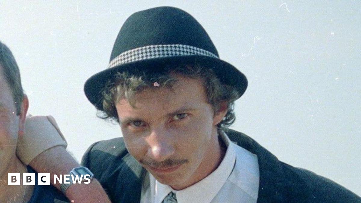 Patrick Murray wearing a black suit and fedora. He has wavy hair and a moustache.