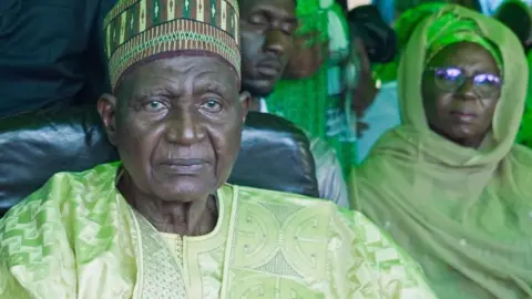Michel Mvondo / BBC Bello Bouba Maigari sits at a campaign rally wearing a green boubou - behind him sits a woman also wearing green.