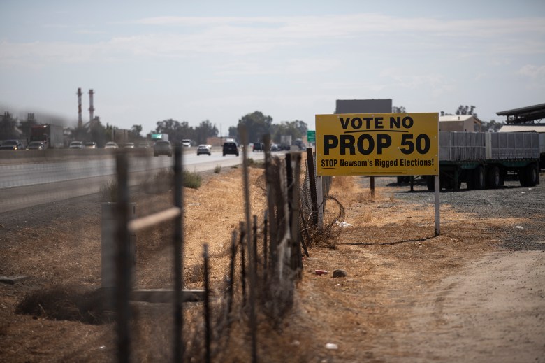 A political sign saying “No on Prop 50” posted on a grassy area near a busy highway along a chain-linked fence on a bright sunny day.