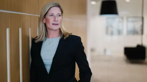 Hill Dickinson Sarah Emerson is a corporate photo. She has blonde hair and is wearing a slick black suit with a grey top and a necklace. We can see she is an office somewhere but the background is blurred.