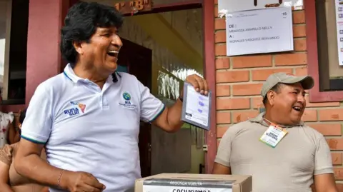 Reuters Morales laughs as he holds up his ballot paper, about to put it into the ballot box. An election official is standing behind him, also laughing - they're both looking off to the side at something.