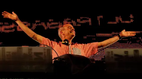 Getty Images A man wearing a red and white checked shirt, with headphones draped around his neck, stands behind a DJ booth, arms outstretched.