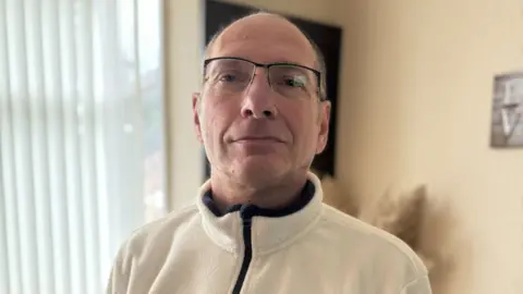 BBC A man stands looking into the camera. He is balding and is wearing glasses and a white fleece top with a zip. Behind him is a beige wall and window with blinds.