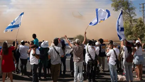AFP via Getty Images Dozens of people stand on the side of the road. They are waving Israeli flags and there is a large dust cloud in front of them. 