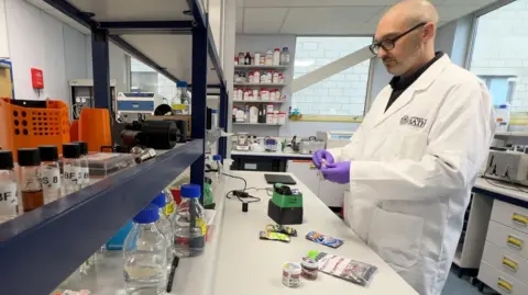 Chris Pudney is wearing a white lab coat, purple rubber gloves and black rimmed galsses. He is stood at a desk in a lab.