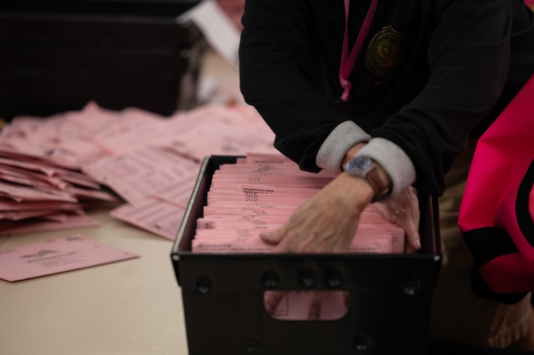 A close-up view of a person's hands sifting through a small black basket filled with pink mail-in ballot envelopes. A stack of pink mail-in ballot envelopes can be seen laying on the table in the background.