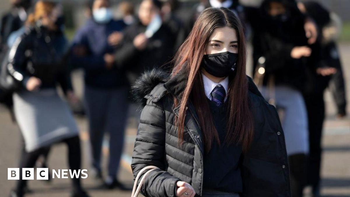 A student in a mask and long brown hair stands facing slightly away from the camera. She is holding a bag and behind her are other students wearing masks.