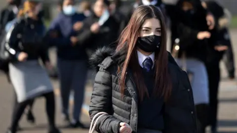 Getty Images A student in a mask and long brown hair stands facing slightly away from the camera. She is holding a bag and behind her are other students wearing masks. 