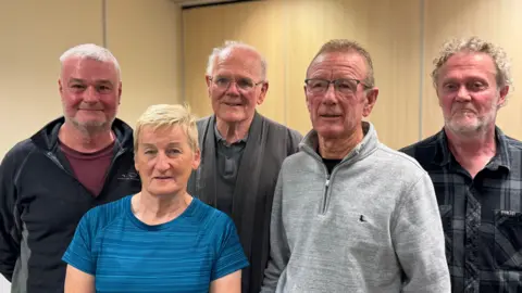 A group of five people stand looking into the camera. 

One man with thinning white hair has short grey stubble and is wearing a black jacket with a red t-shirt. Beside him is another man with short white hair who is smiling. He is wearing metal rimmed glasses, a grey jacket and a grey polo shirt. There is a woman with short blonde hair wearing a blue striped top and gold hoop earrings. Another man with short hair is wearing a grey jumper with a zip and a black top underneath. He is wearing grey framed glasses. And the fourth man on the right side of the image has curly grey hair is looking into the camera. He has a short white beard and is wearing a grey check button up shirt. 