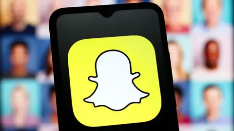 Getty Images Snapchat yellow and white "ghost" logo is displayed on a black smartphone screen, with blurred images of people's faces in the background.