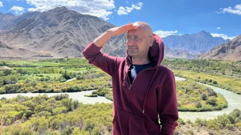 A bespectacled man looks ahead, standing in front of a winding river in a mountain valley