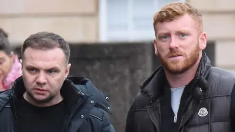 Spindrift Eddie Lyons Jr (left) and Ross Monaghan (right) wearing dark clothing. Monaghan has red hair and is looking right at the camera. Lyons Jnr is looking off camera and has dark hair.
