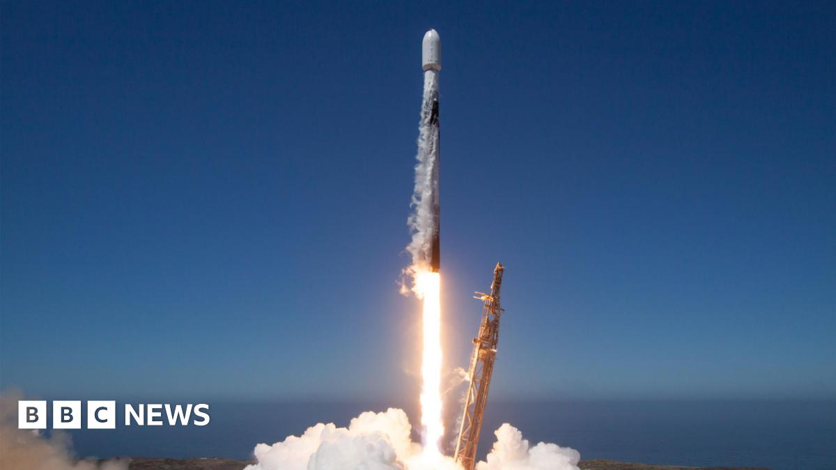The exterior of the air force base. It's a tall, grey structure surrounded by small huts and barbed-wire.