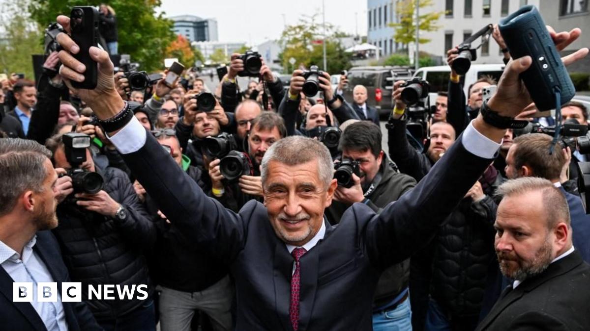 Andrej Babis dressed in a dark suit with his hands in the air in celebration, surrounded by journalists taking photos of him.