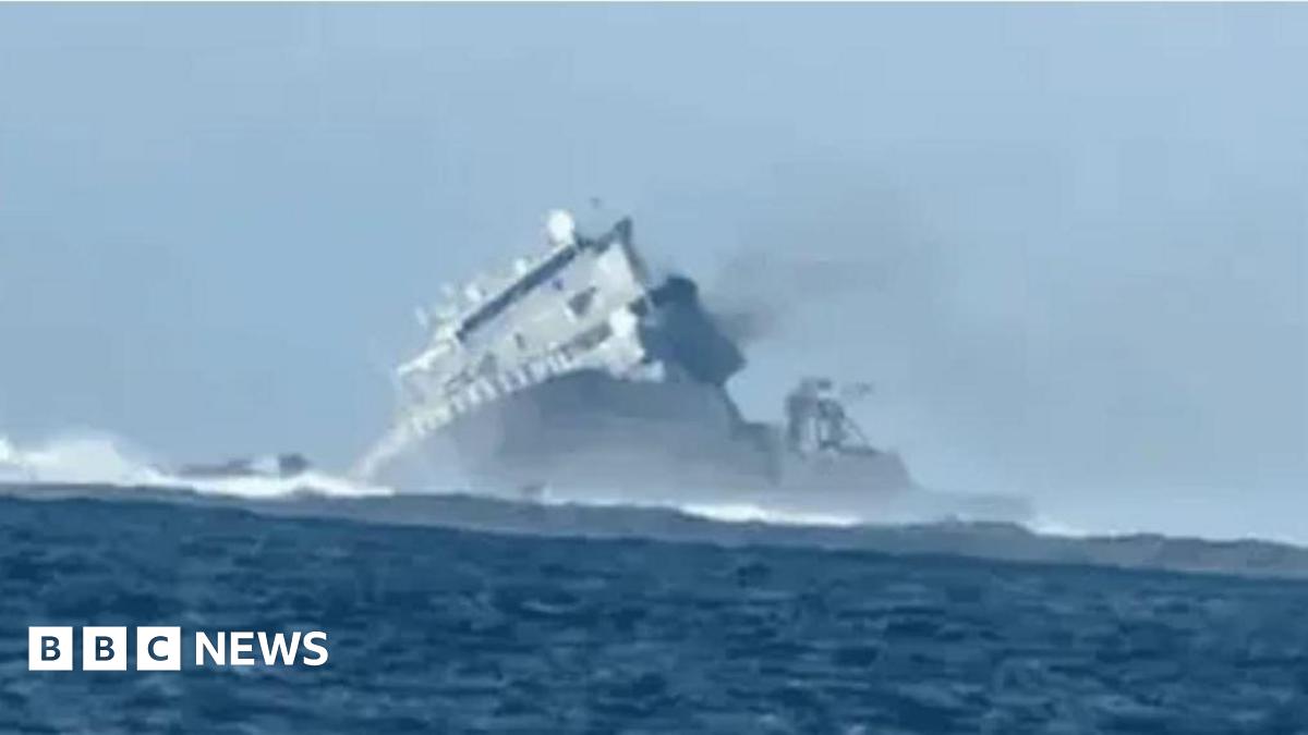 Kemi Badenoch wearing a white jacket and white t-shirt is pictured waving to the camera. There is a union jack pictured to the left.