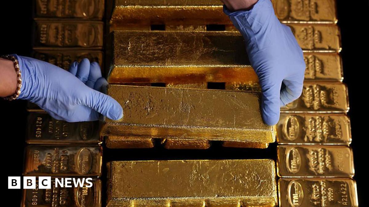 A pair of light blue latex-gloved hands place a gold bar on top of pile of other gold bars. The person has a gold bracelet on their right wrist.