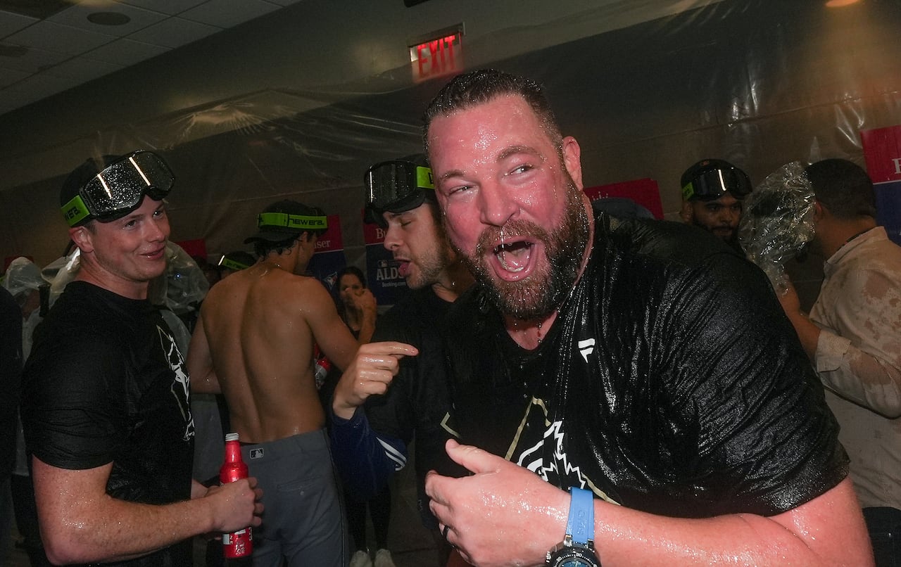 A man with a goatee wearing a black t-shirt yells happily as he is soaked in champagne, surrounded by baseball players wearing goggles and holding champagne and beer bottles in a room draped in plastic sheeting.