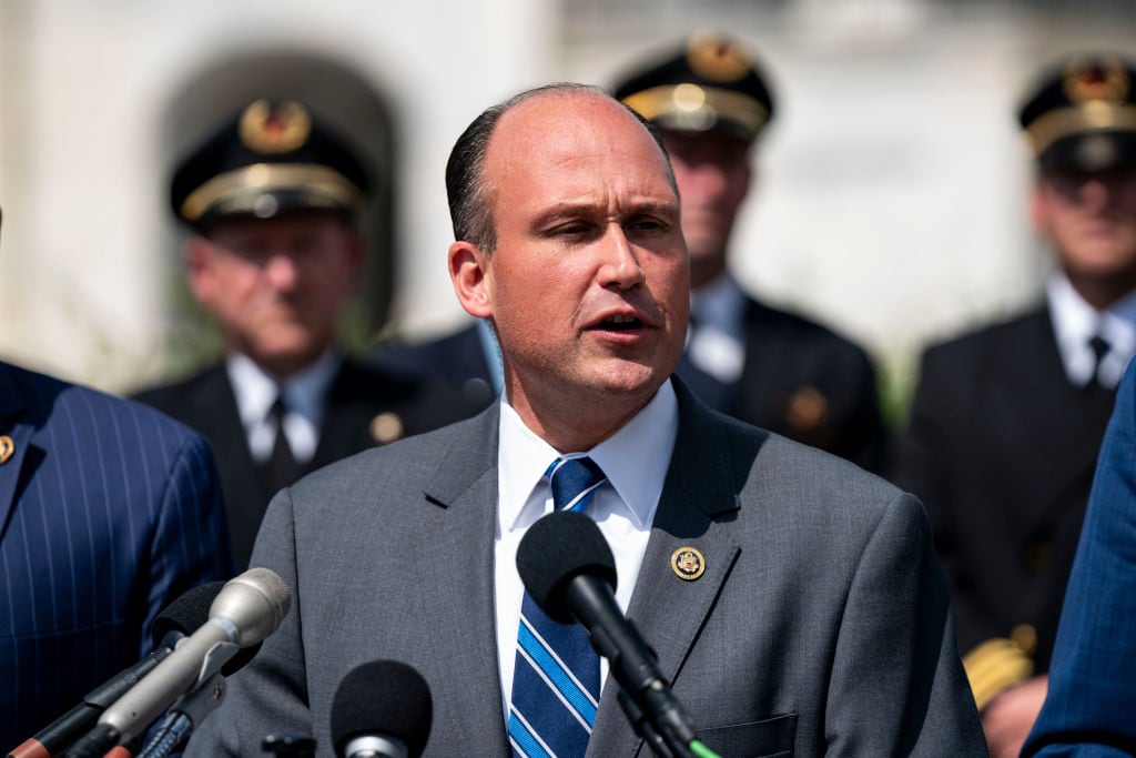 A man speaks outside with military personnel behind him.
