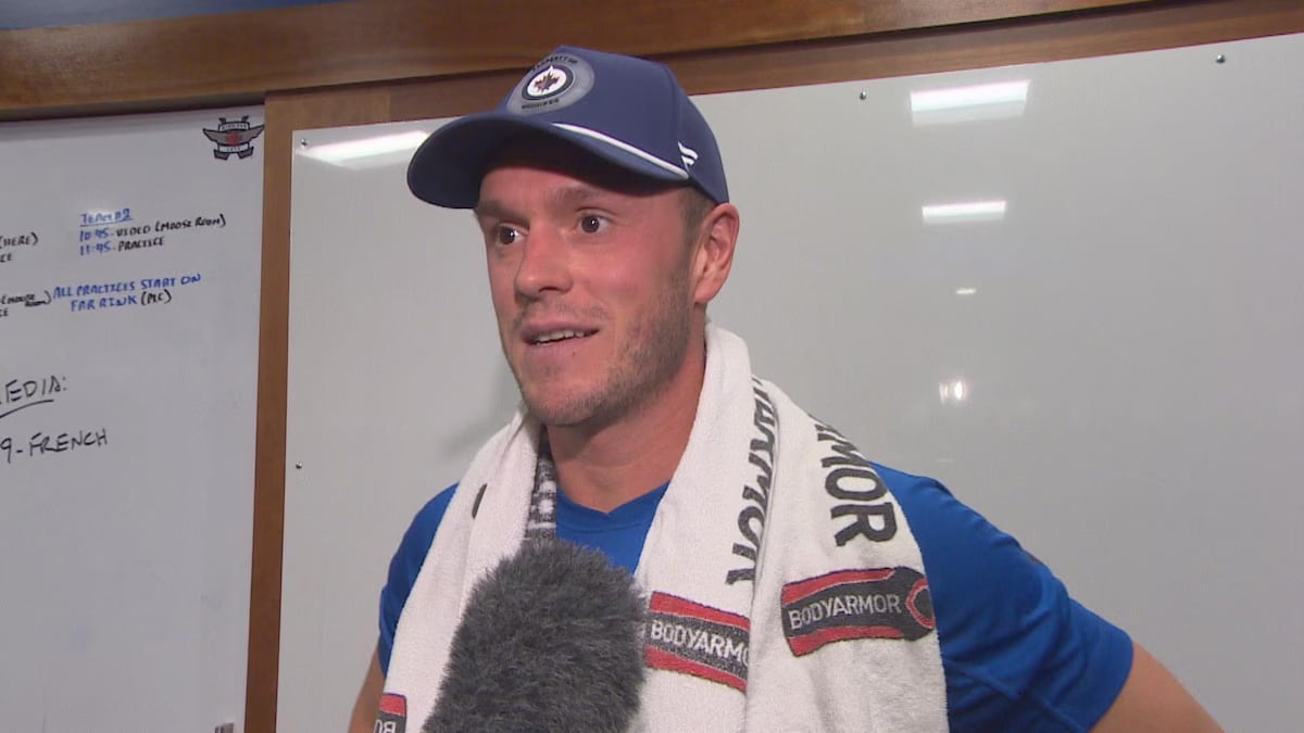 A man wearing a Winnipeg Jets ball cap with a towel draped over his shoulders speaks into a microphone.